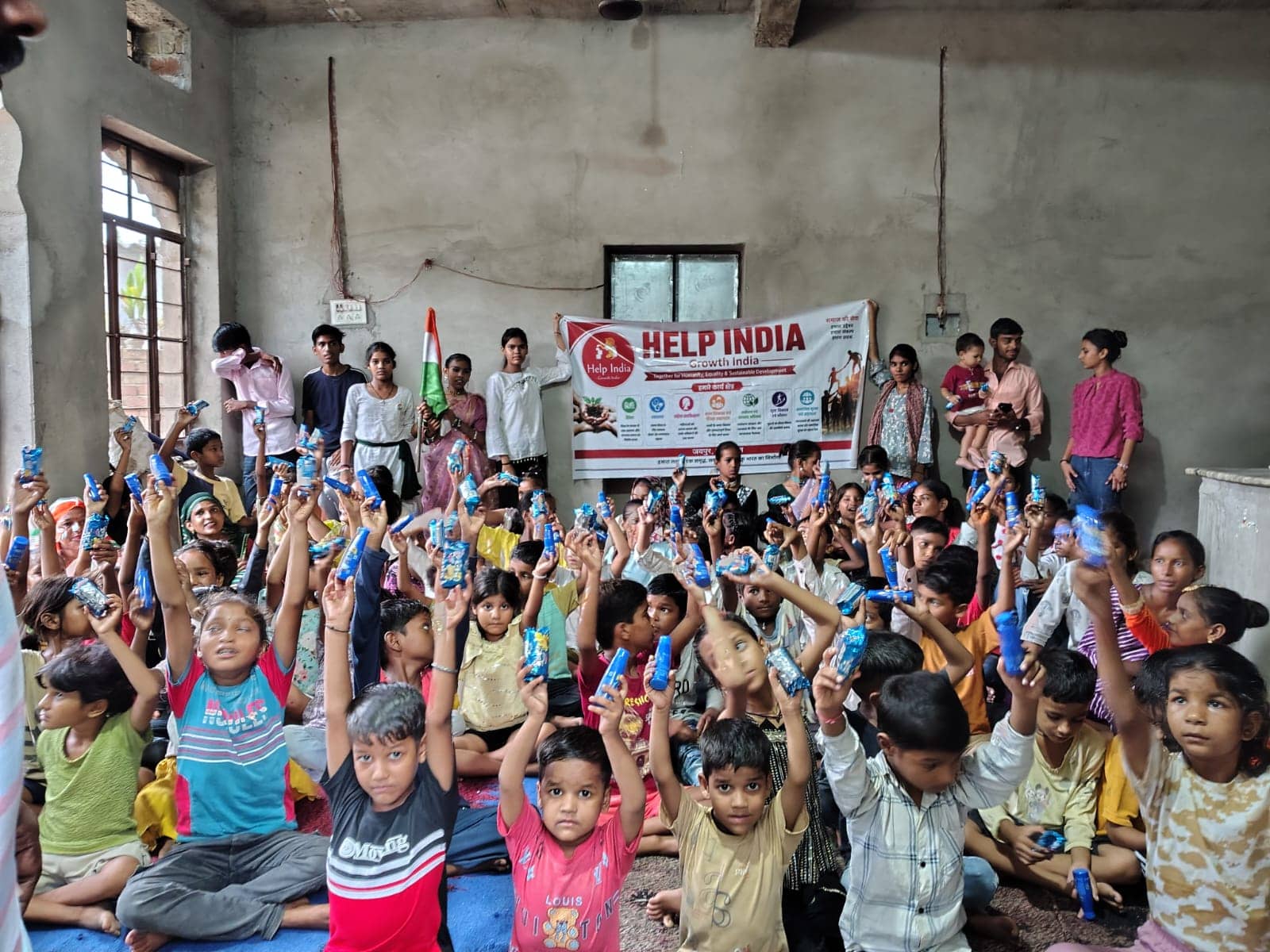 Children seated on the floor raise small packets in the air, with volunteers and the Help India Growth India banner behind them.