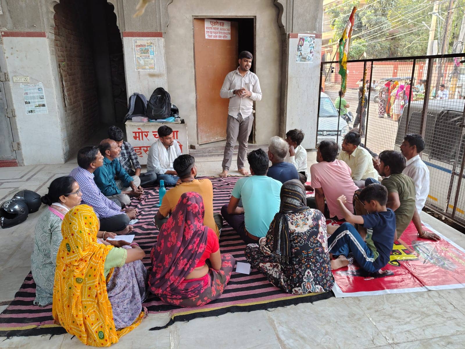 A man stands and speaks to women and men seated on a striped durrie in a shaded courtyard.