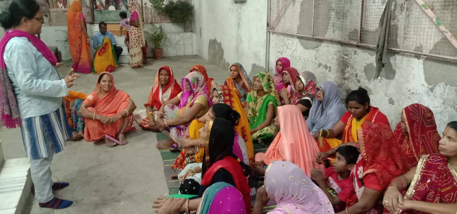 A woman in a light blue kurta and pink scarf stands and speaks to women seated on mats in a courtyard.