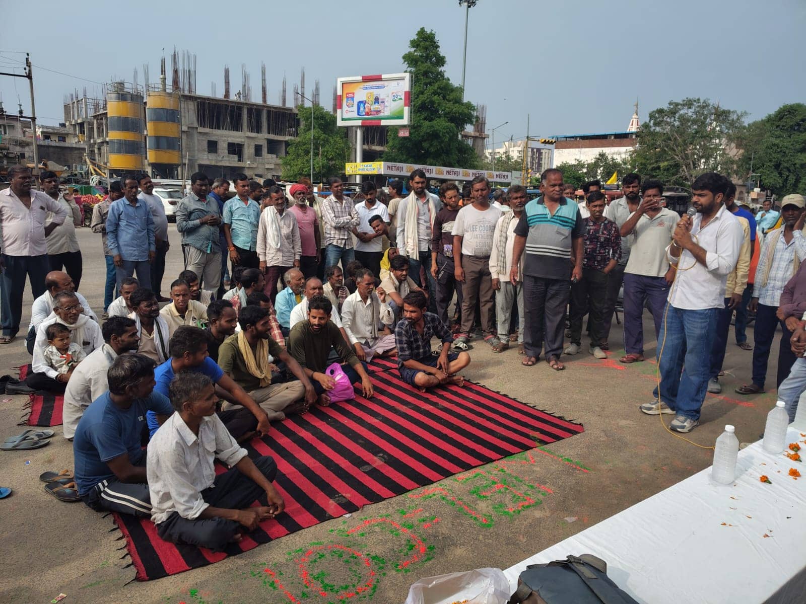 A wider view of an open-air gathering of workers listening to a speaker, with buildings under construction in the background.